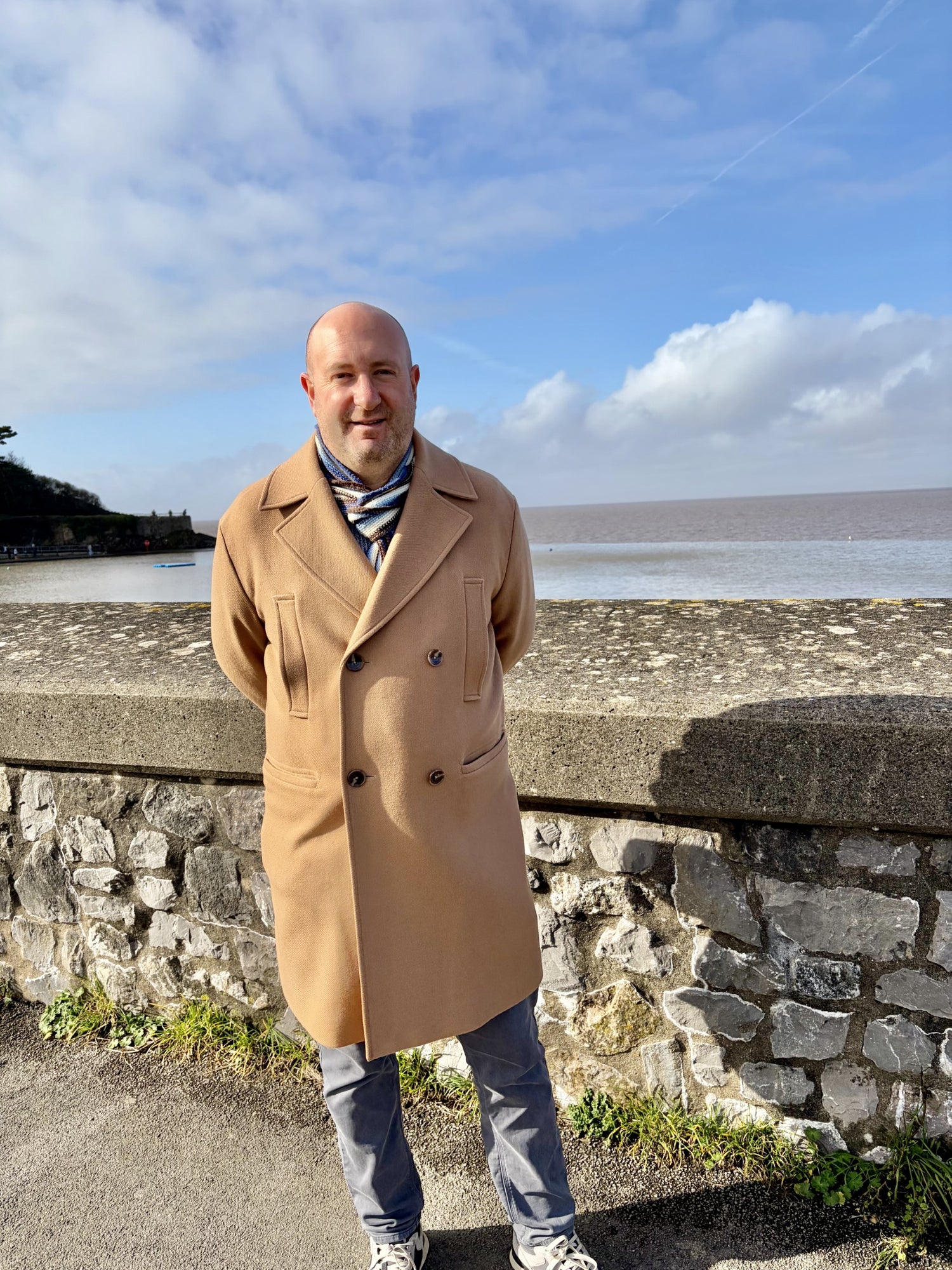 Daniel Boden (founder) in a beige coat standing by a stone wall with a scenic background